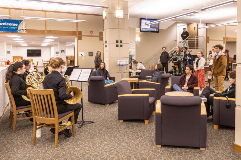 A group of people sit and stand while four horn players perform on the first floor of the library.