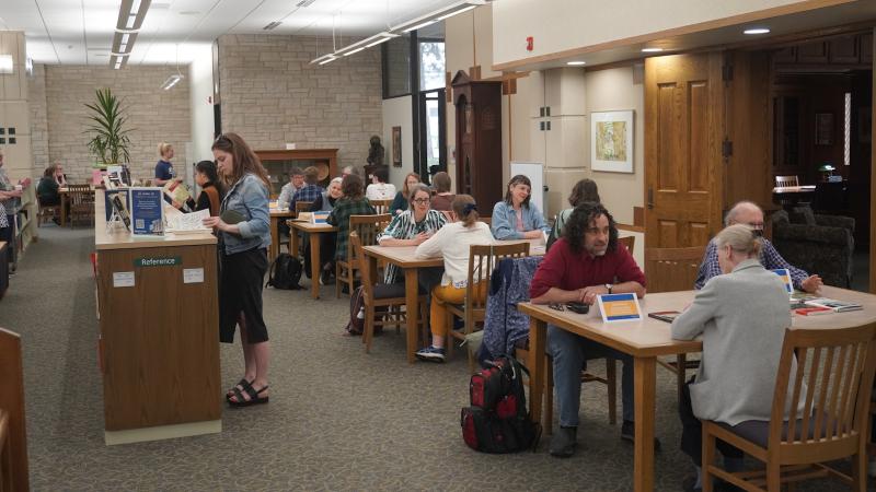 First floor of the library with people sitting at tables and standing at shelves during an author event.