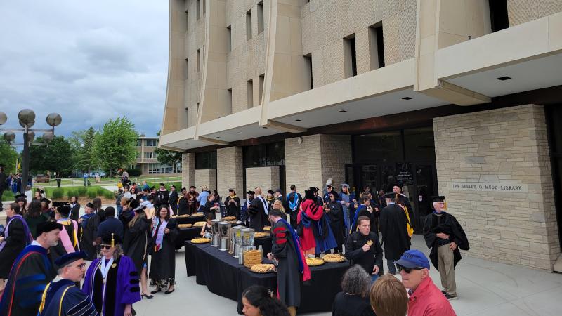 Crowd of faculty and students in graduation regalia as well as friends, family, and catering staff, gathered in front of the Mudd Library. Tables with clear water dispensers and trays of cookies are set up.