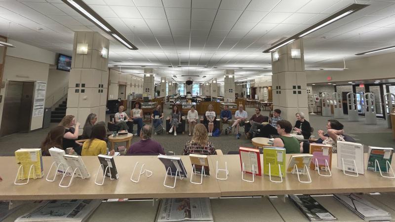 A group of people in the library sitting on chairs in a circle holding and talking about books. A display of books on stands is in the foreground.