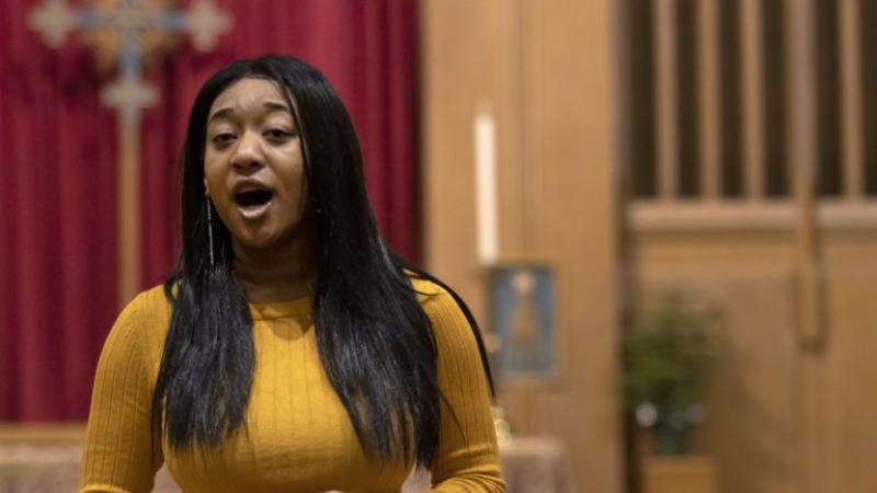 Hannah Jones sings in a church, wearing a yellow sweater.