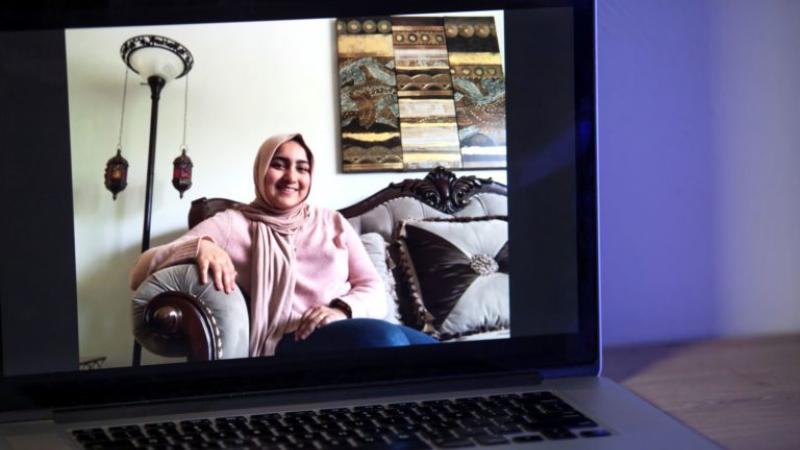 A laptop screen shows Ghania Imran, wearing a pink sweater and sitting on a grey couch.