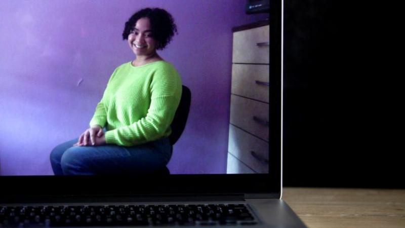 Sabrina Salas, sitting in a chair and wearing a green sweater, is shown on the screen of a laptop.