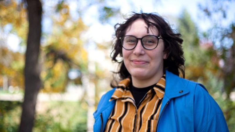 Dani Massey, wearing a blue jacket and yellow and black striped shirt, smiles at the camera.