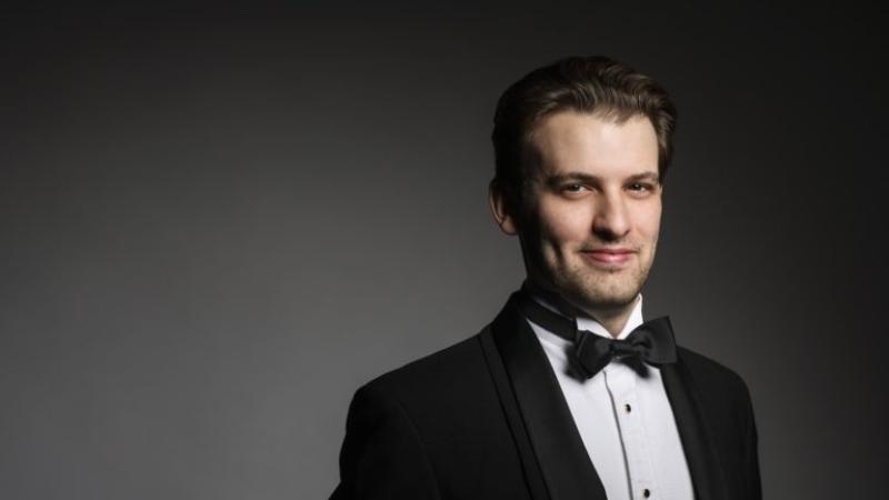 Evan Bravos, wearing a tuxedo, smiles at the camera against a black background.