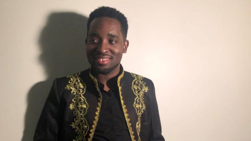 Louric Rankine, wearing a black and gold jacket, stands in front of a white wall and smiles.