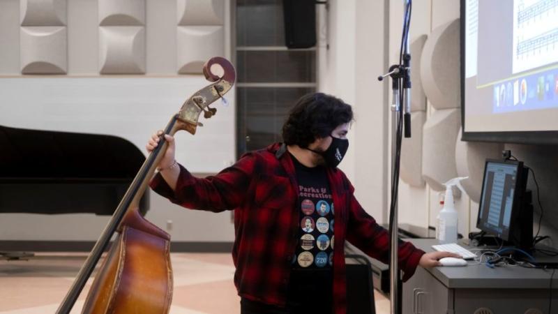 Student holding a double bass in one hand and working on a computer with the other