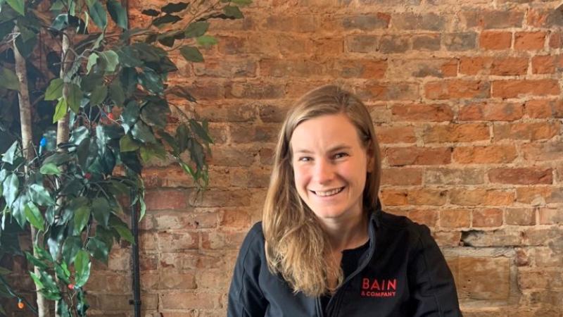 Emily Muhs, wearing a "BAIN" jacket, smiles at the camera against a brick wall.