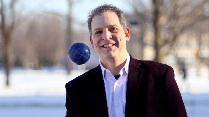 David Gerard, wearing a black suit jacket, throws a blue ball in the air.