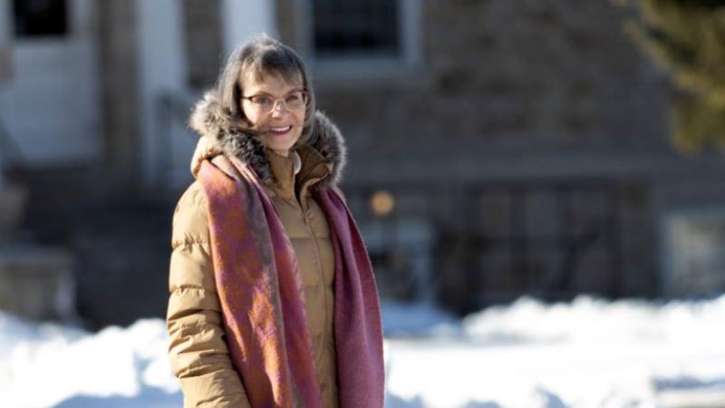 Eilene Hoft-March, wearing a tan coat and pink scarf, smiles at the camera.
