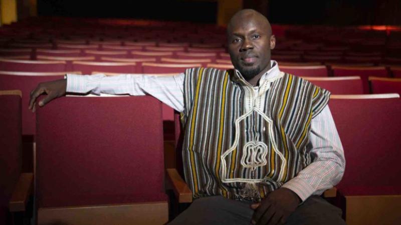 Patrick Adu sits in a red chair in a theater, wearing a striped vest.