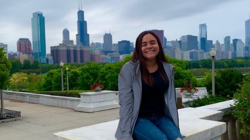 student Stephany Pichola sitting on a stone railing with the background of the Milwaukee skyline 