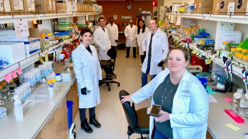 Seven people, all wearing white lab coats, stand in a room full of laboratory equipment.