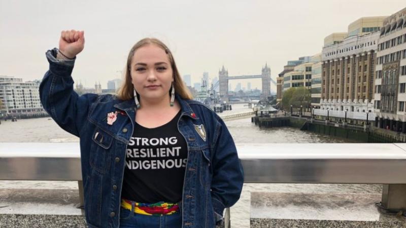 Shelby Siebers, wearing a shirt with the words, "Strong, Resilient, Indigenous," raises her fist in the air.