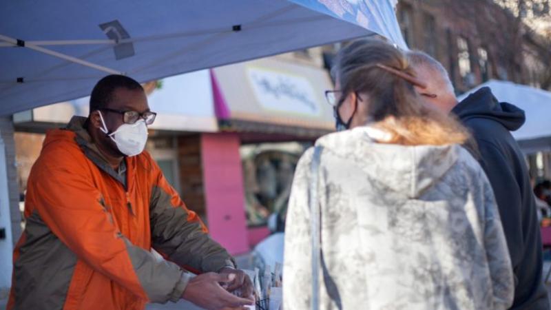 Yaw Asare, wearing an N95 mask and orange coat, interacts with a customer at his Farmers Market booth.