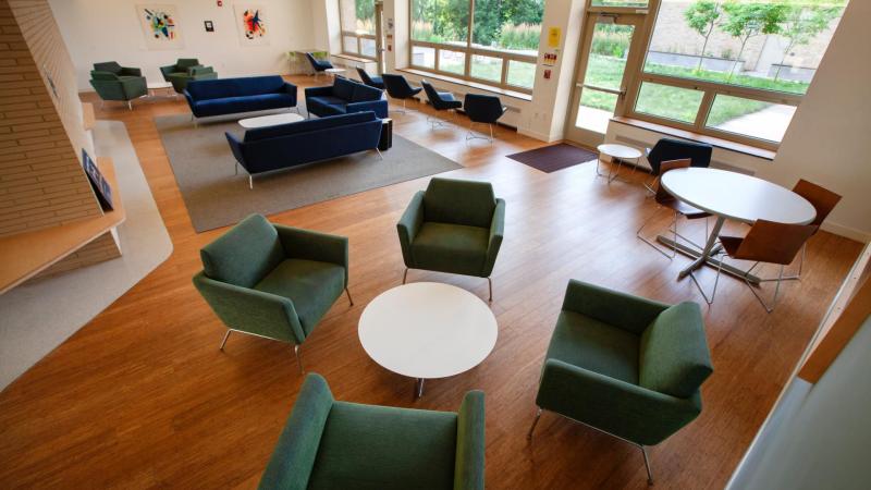 open common area in dorm with large windows, green and blue chairs and sofas circling white tables
