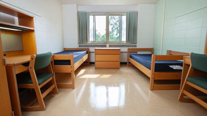 large window over wooden drawer between two XL twin beds and a green chair at a desk