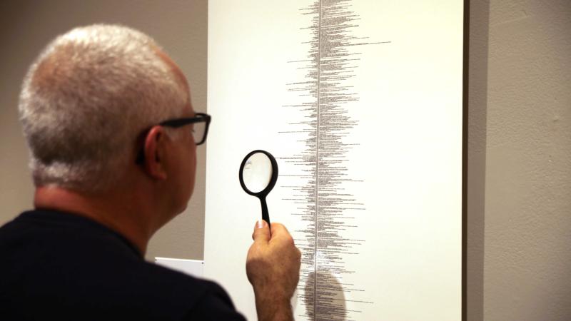 A man in a black t-shirt seen from behind as he holds a magnifying glass up to a drawing with tiny text on a long white piece of paper.