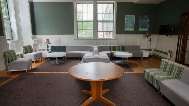 Below large window are mirrored section sofas around a white table with vintage lamps on either end. A large wooden table in the foreground surrounded by armchairs