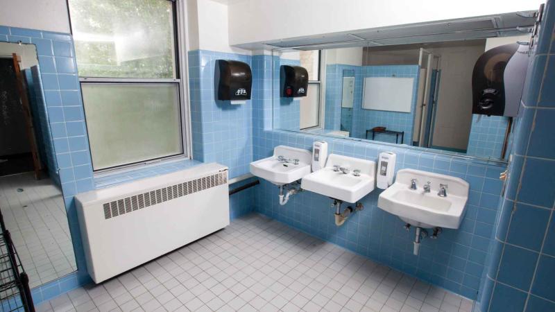 Tiled bathroom with a long mirror over several sinks and reflection of toilet stalls and showers in mirror reflection