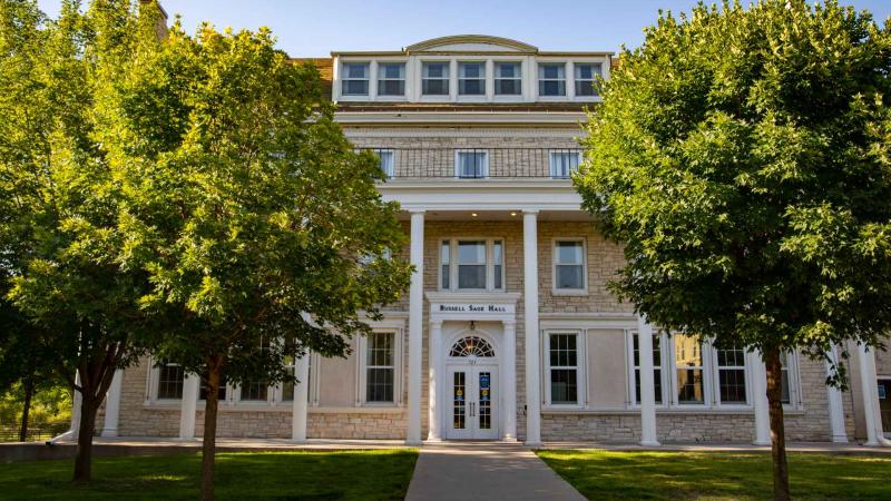 Colonial style building with stately archways and herculean pillars with brick path leading to main door flanked by trees and well-kept lawn