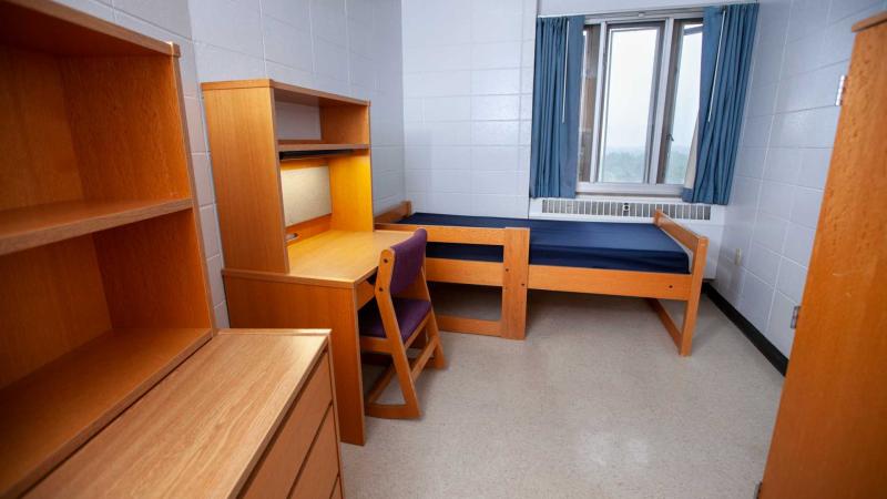 White single dorm room with a small window, one XL twin bed, one desk and drawer made of wood.