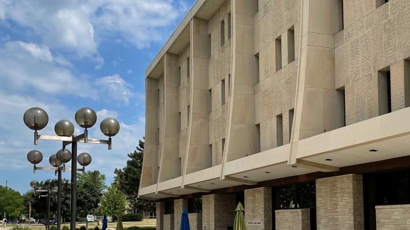 Exterior view of the Mudd Library building, when standing off to the right. Iconic globe light fixtures, green grasses, and a blue sky with fluffy clouds surround the building. Outdoor tables and chairs are located just outside the building entrance.