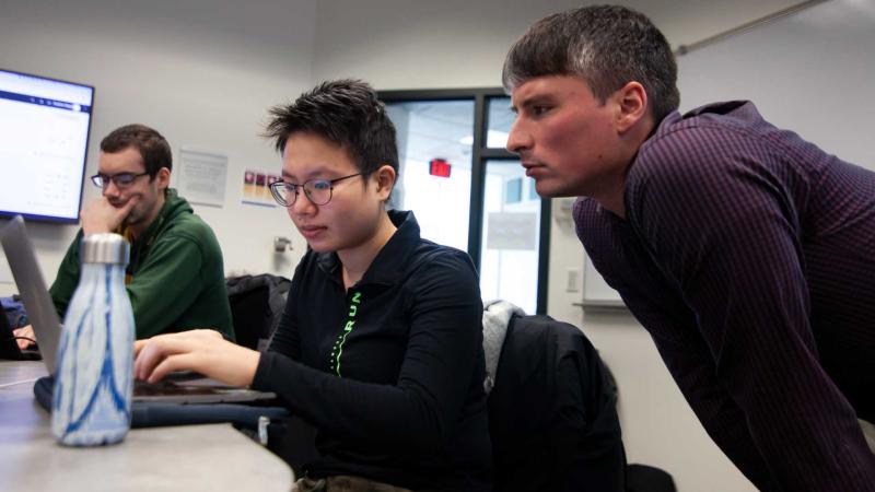 computer science professor helping student on laptop seated at long table during class