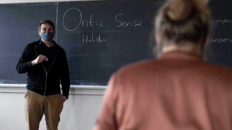 philosophy professor standing in front of blackboard with student listening in foreground