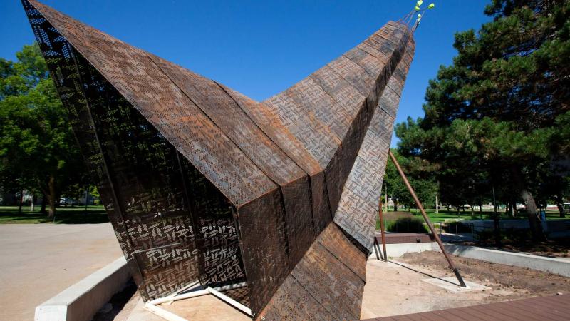 Giant sculpture with perforated and patinaed steel panels and geometric beadwork designs on them, overlap like a crane's feathers.