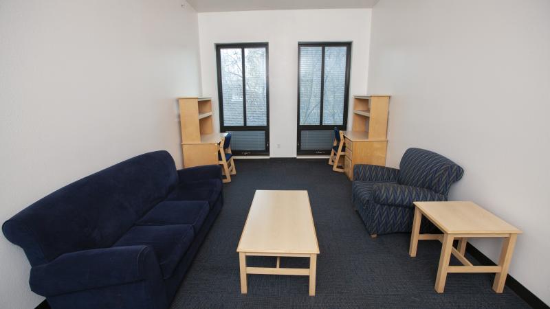 Common living space with two desks and shelves, two long vertical windows, a couch and armchair with a coffee table in between them.