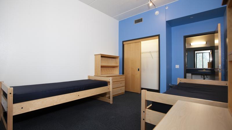 Double room in Hiett Hall with two beds, two dressers and shelves overhead, a closet, and bathroom in the back.