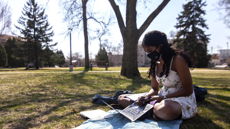 Adya '23 sitting on Main Hall Green reading with a mask on.