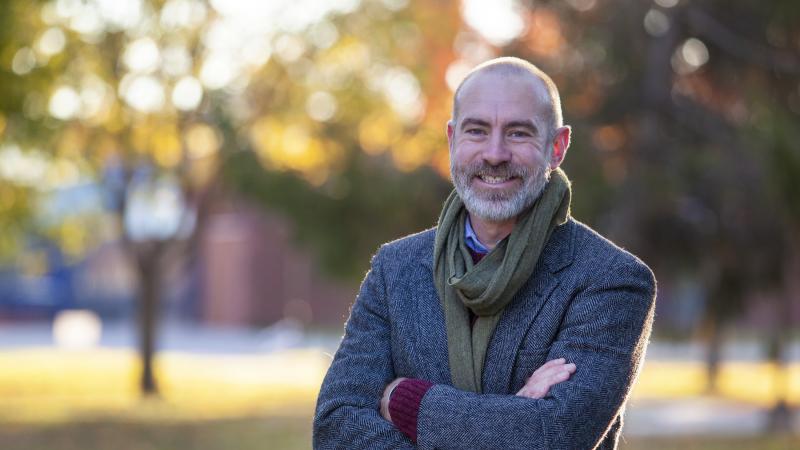 Jason Brozek, wearing a grey tweed blazer, stands on Main Hall Green. 