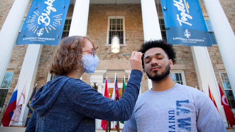 image of a student painting another students face in front of main hall