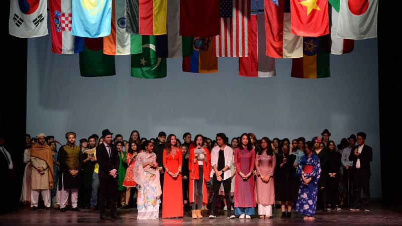 Many students onstage in their ethnic clothing with various country flags hanging down.