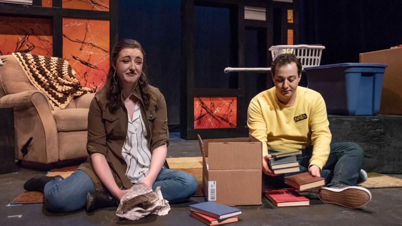 Two actors sitting amongst books with sad expressions on their faces during dress rehearsals for "Buried in Debris."