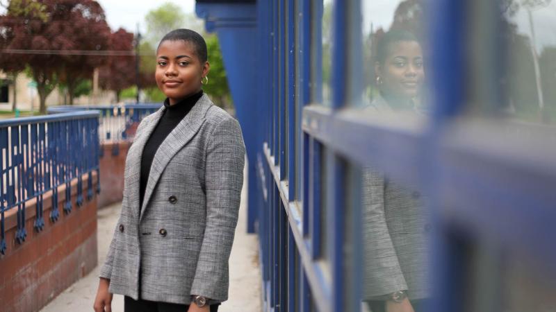 Shania Johnson. wearing a grey suit jacket, stands next to the blue windows of the Wriston Art Center.