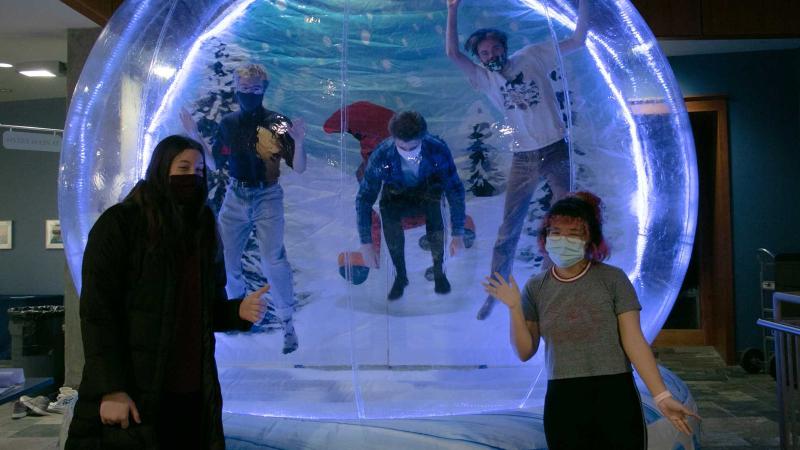 Three students in masks jumping up in large plastic snow globe glowing blue while two students in masks pose outside.