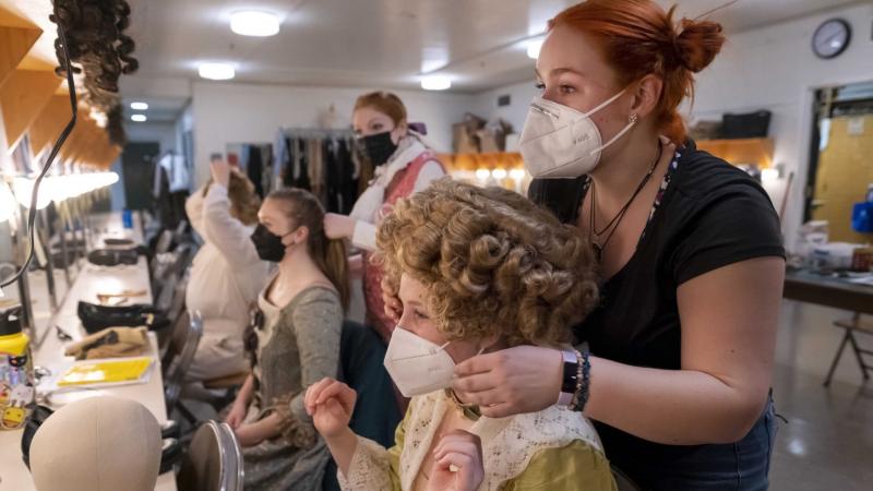 Student actors in victorian era costumes sit in front of their mirrors in the green room while other students do their hair.