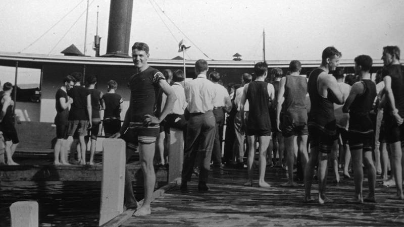 Men in old-fashioned swimsuits stand on dock near the Fox River with a bridge in the background