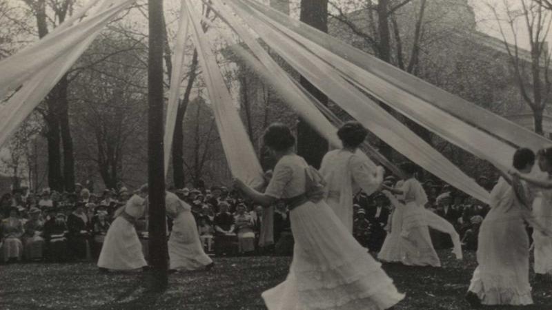 Women wearing white dresses holding white cloth tied to a tall pole while walking around it.