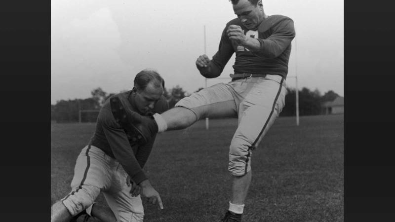 Two student in football uniforms, the placekicker is kneeling on the grass, while the kicker has just kicked the ball