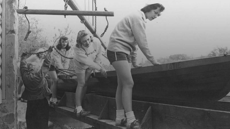 Women standing on long row boat as it hangs over the Fox River