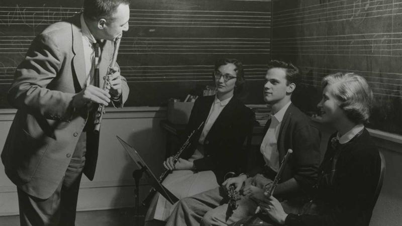 Professor standing playing the flute while three students sit with their flutes in hand, listening 