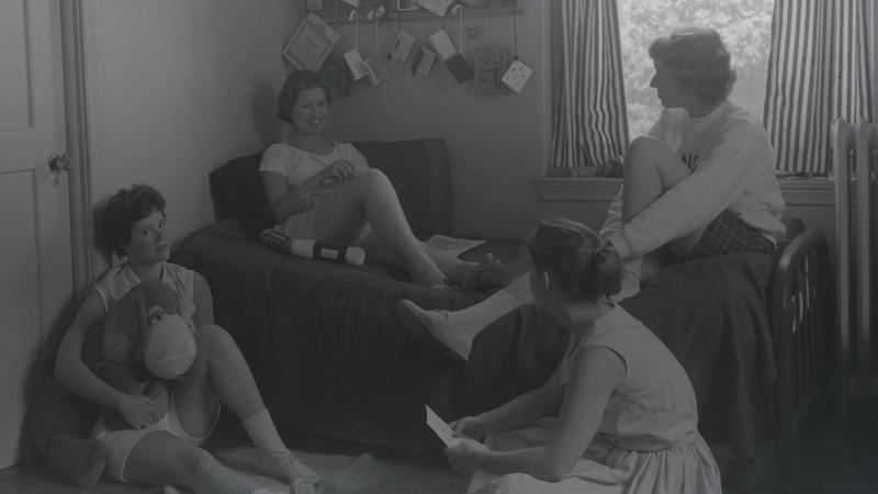 Two students sit on a lounge sofa in front of a window while two others sit on the ground, chatting