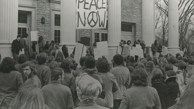 Crowd of students gather in front of Main Hall to protest. A sign that says, "peace now" hands in between two pillars of the building.