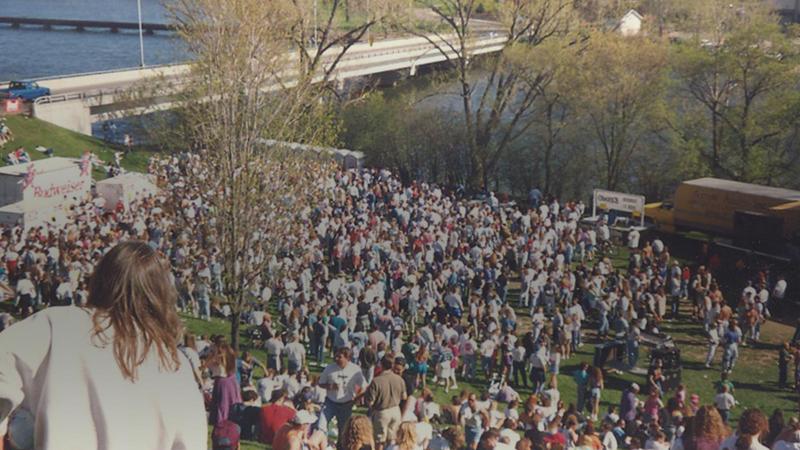 A large crowd of students gather on campus. The Fox River and bridge can be seen in the background