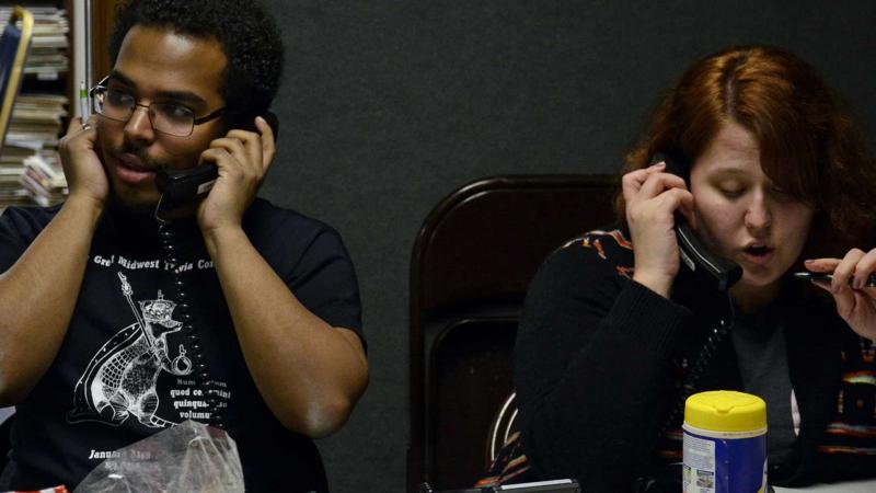 Two students seated at a table speaking into hand-held phones