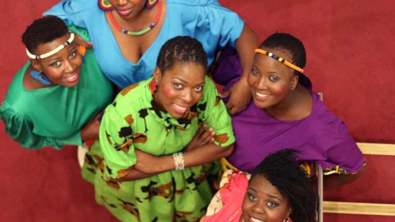 Performers of Nobuntu wearing colorful dresses and looking up at the camera standing on red carpet.